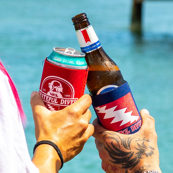 two people cheersing their drinks in Grateful Diver Can Koozies with water in the background