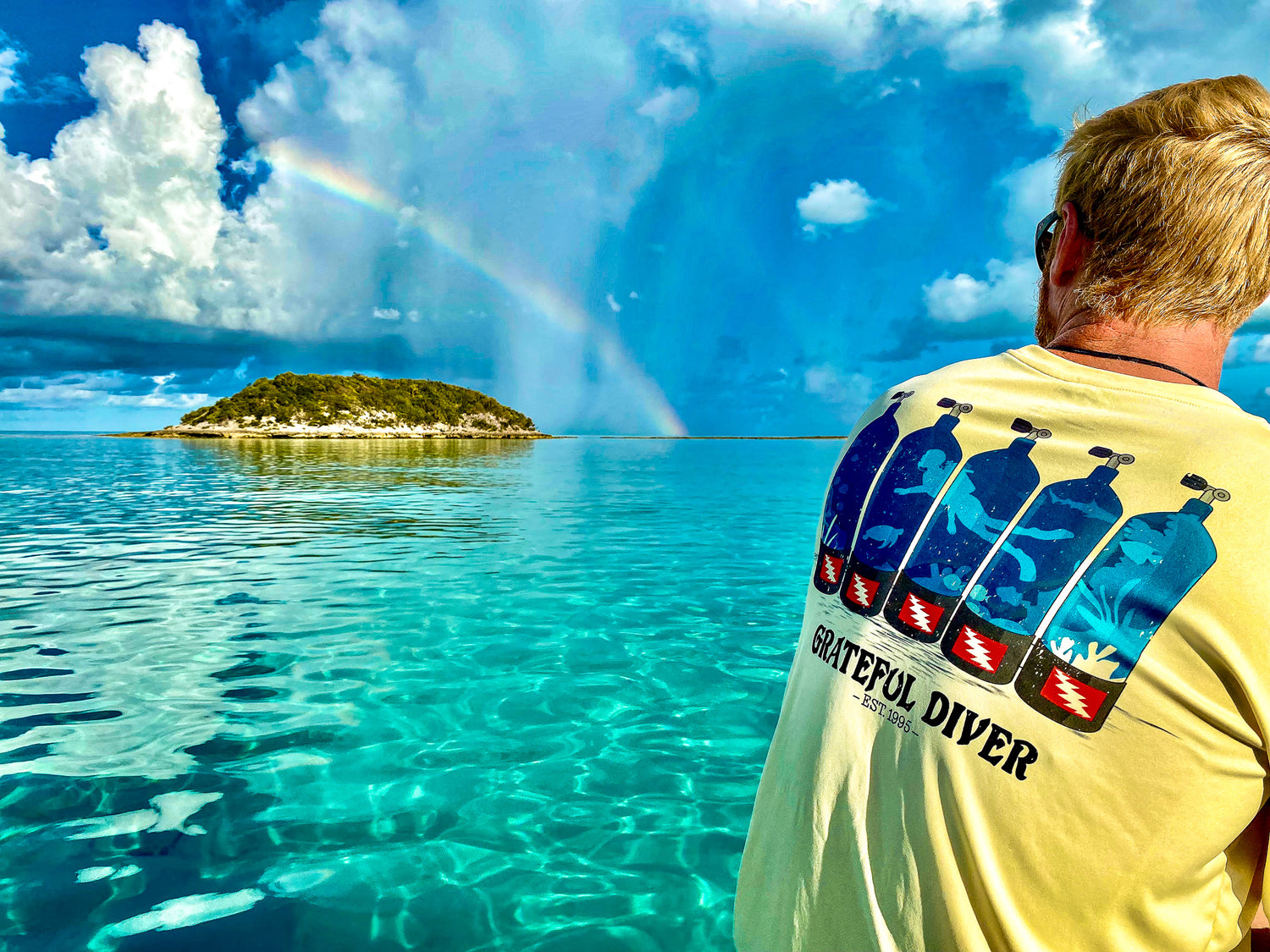 man in pale yellow Grateful Diver Dive Tanks UV Shirt looking across ocean at rainbow over an island