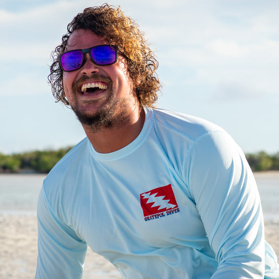 smiling man in sunglasses sitting on sandbar wearing a Grateful Diver UV Shirt