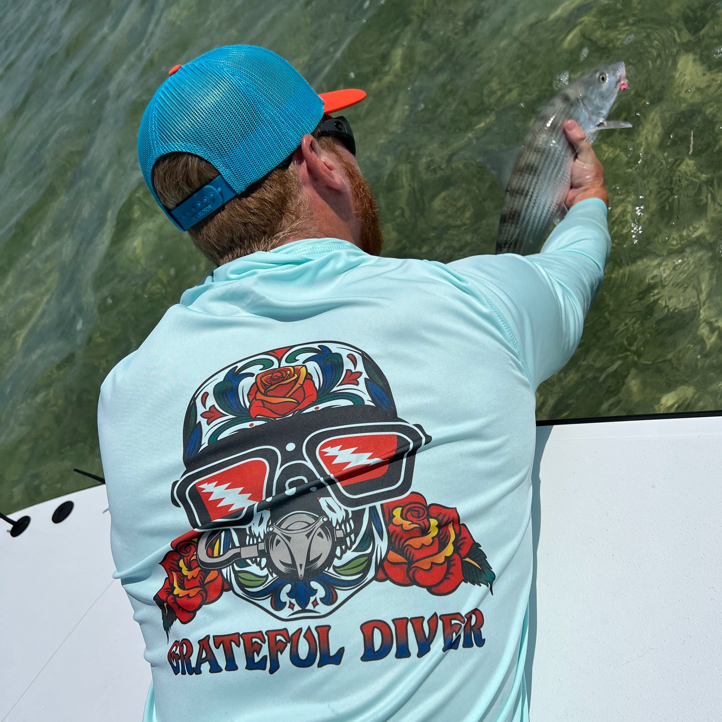 Grateful Diver Sugar Skull UV Shirt Hoodie in arctic blue on model showing back while holding a fish off the side of a boat