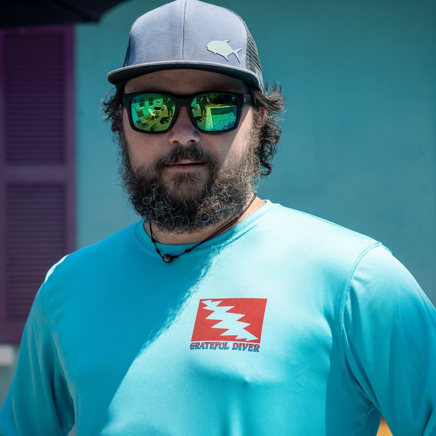 Grateful Diver Reef Diver UV Shirt in water blue on man in a hat and sunglasses showing front lightning bolt logo with the building in the background