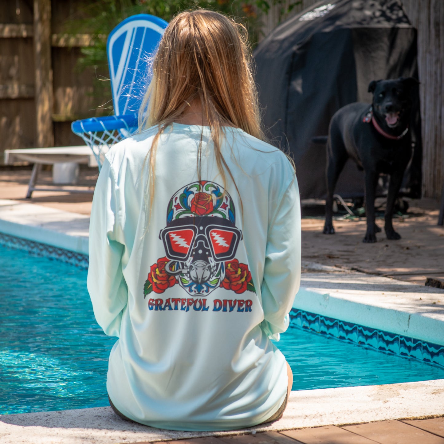 Grateful Diver Sugar Skull UV Shirt in arctic blue showing back on woman sitting by dog & pool