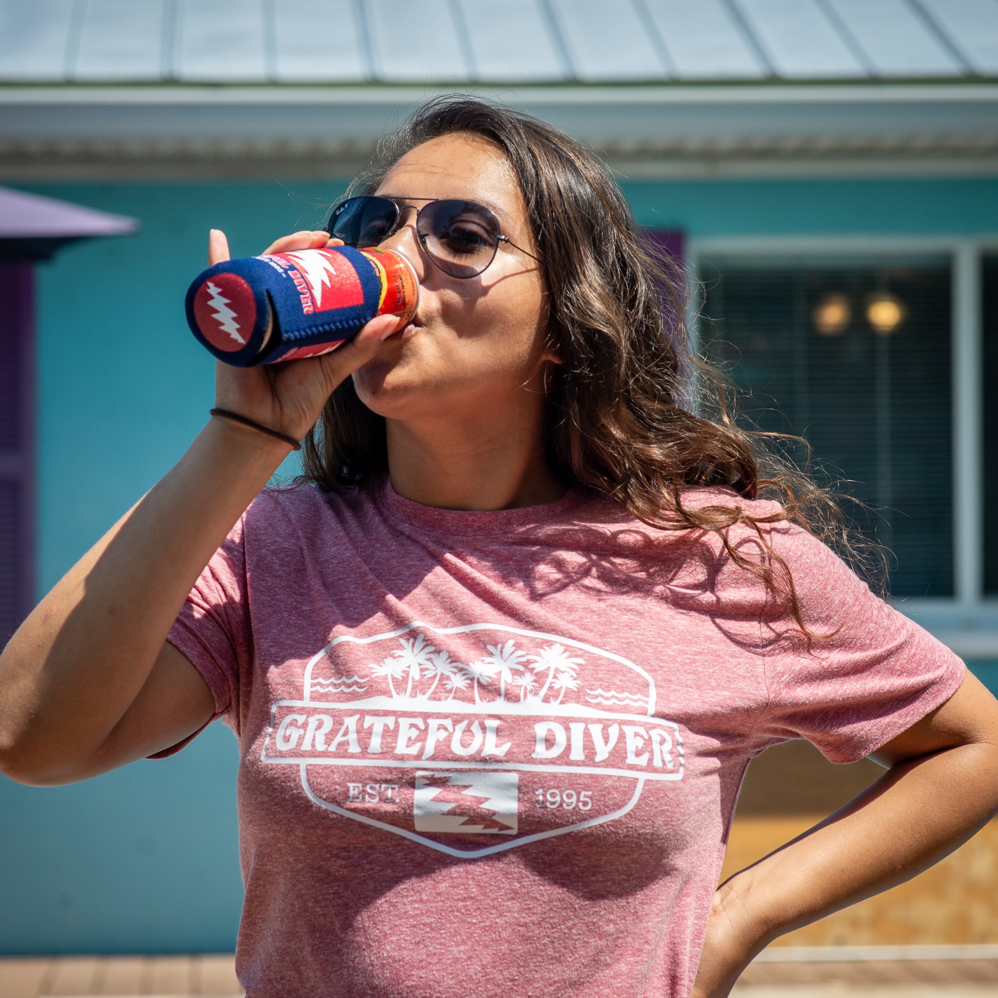 Grateful Diver Palm Island T-shirt in red on woman taking a drink from a Grateful Diver can koozie in front of building